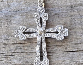 May include: A silver-colored pendant in the shape of a cross, encrusted with small, sparkling stones. The cross has a detailed design with decorative elements at the ends of the arms and a loop at the top for a chain. The pendant is displayed on a wooden surface.