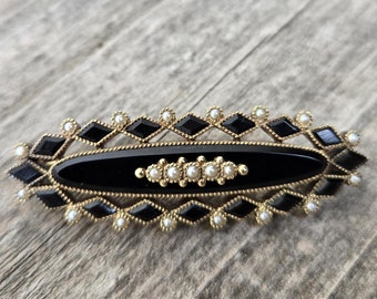 May include: An oval brooch featuring a black central stone, framed by gold-coloured metal and black diamond-shaped details. Small, white pearl-like beads are set within the design. The brooch is displayed on a weathered wooden surface.