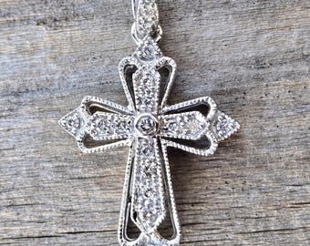 May include: A silver-colored pendant in the shape of a cross, encrusted with small, sparkling gemstones. The cross has an ornate, openwork design with a loop at the top for a chain. The pendant is displayed against a weathered wooden surface.