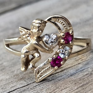 May include: A gold-toned ring featuring a cherub holding a heart-shaped design. The heart is adorned with a white gemstone and two red gemstones. The ring has a split shank design and is set against a wooden surface.