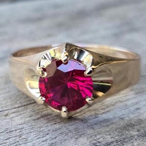 May include: A gold ring featuring a large, round, vibrant red gemstone. The ring has a classic design with a smooth, polished band and decorative accents around the gemstone. The gemstone is the focal point, catching the light and adding a touch of elegance.