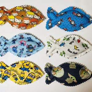 May include: Six fabric fish-shaped toys with pinked edges, featuring various character prints including Scooby-Doo, The Cat in the Hat, Stitch, Dr. Seuss text, Minions, and Grogu on blue, yellow, white, and dark gray backgrounds.