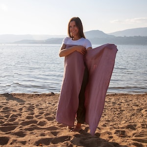 May include: A woman stands on a sandy beach, holding a long, dusty rose-colored linen wrap. The wrap is draped around her, with the ends flowing in the breeze. The background features a lake, mountains, and a clear sky.