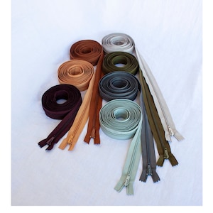 May include: Assortment of zipper tapes in various colours, including brown, beige, green, grey, and blue. The zippers are rolled up and the ends are unzipped, showing the zipper teeth.