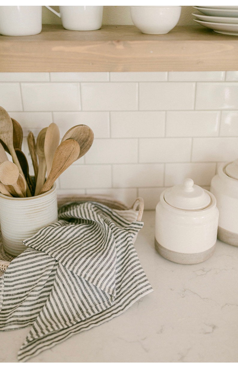 Pu&ograve; includere: Scena di cucina con cucchiai di legno in un portautensili di ceramica bianca. Un canovaccio a righe bianche e nere poggia su un vassoio intrecciato. Sono visibili anche due barattoli di ceramica bianca con coperchi.