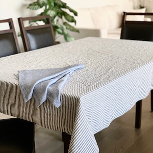May include: A white linen tablecloth with thin blue stripes, folded in half and placed on a wooden table. The tablecloth is slightly creased and has a casual, relaxed look.