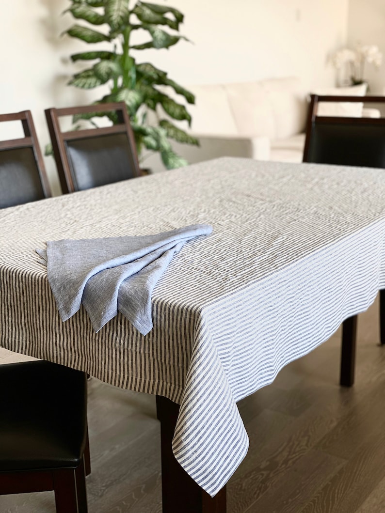 May include: A white linen tablecloth with thin blue stripes, folded in half and placed on a wooden table. The tablecloth is slightly creased and has a casual, relaxed look.