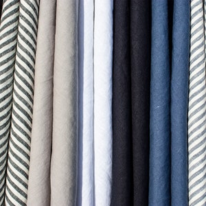Pu&ograve; includere: Una collezione di tessuti di lino piegati in vari colori. I colori includono nero, grigio, bianco, blu navy e blu. Un tessuto presenta un motivo a righe bianche e grigie. I tessuti sono disposti verticalmente, creando un'esposizione visivamente accattivante di trame e tonalit&agrave;.