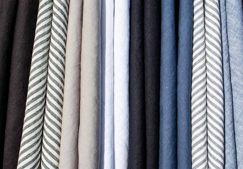 Pu&ograve; includere: Una collezione di tessuti di lino piegati in vari colori. I colori includono nero, grigio, bianco, blu navy e blu. Un tessuto presenta un motivo a righe bianche e grigie. I tessuti sono disposti verticalmente, creando un'esposizione visivamente accattivante di trame e tonalit&agrave;.