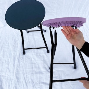May include: Two folding stools with black metal frames. One stool has a navy blue seat, and the other has a lavender seat with a ruffled edge. The stools are on a white background.
