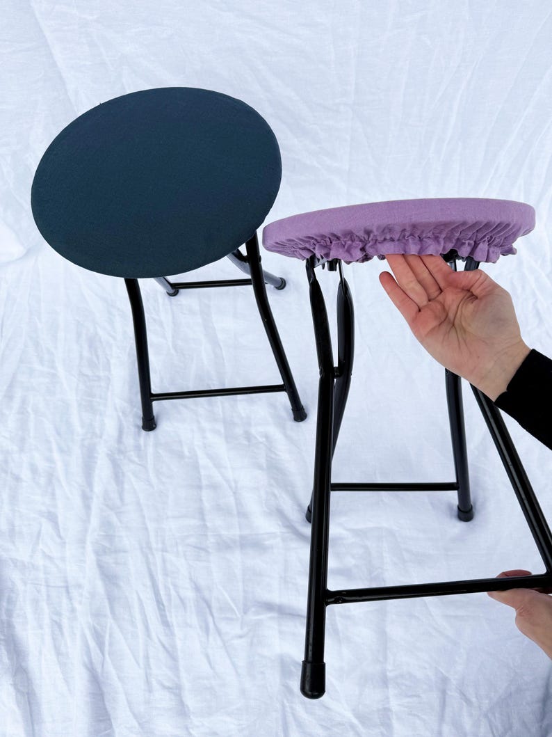 May include: Two folding stools with black metal frames. One stool has a navy blue seat, and the other has a lavender seat with a ruffled edge. The stools are on a white background.