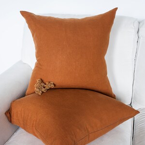 May include: Two brown linen throw pillows on a white sofa. The top pillow is slightly larger than the bottom pillow.
