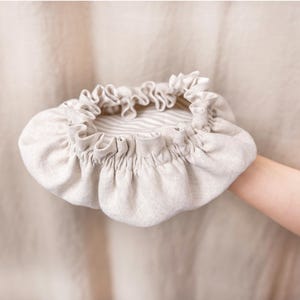 May include: A beige linen bonnet with a ruffled edge and a striped interior. The bonnet is held up by a person's arm. The fabric has a soft, natural texture, and the design is simple and elegant.