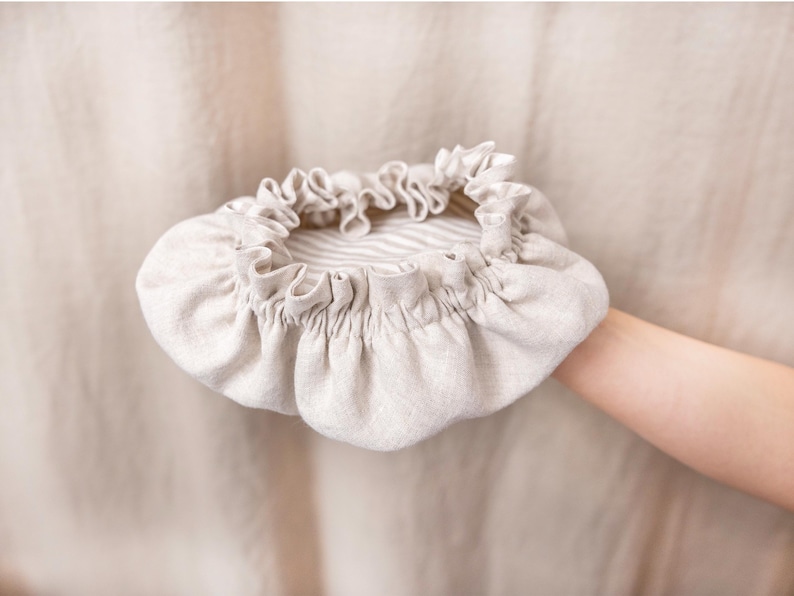 May include: A beige linen bonnet with a ruffled edge and a striped interior. The bonnet is held up by a person's arm. The fabric has a soft, natural texture, and the design is simple and elegant.