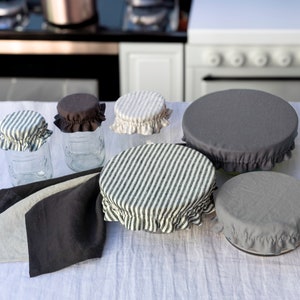 Peut inclure: Un ensemble de six housses alimentaires réutilisables en tissu de différentes tailles et motifs. Les housses sont faites d'un tissu doux et naturel et sont conçues pour s'adapter sur des bols, des bocaux et d'autres récipients afin de garder les aliments frais. Les housses sont présentées sur une nappe blanche avec deux serviettes en tissu.