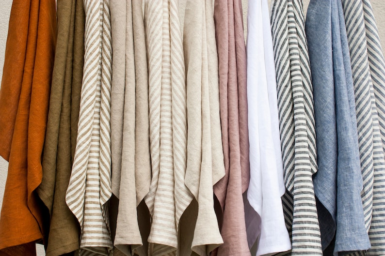 Pu&ograve; includere: Una collezione di tessuti di lino in vari colori, tra cui ruggine, verde oliva, beige, rosa, bianco e blu. Alcuni tessuti presentano motivi a righe. I tessuti sono appesi, mostrando la loro consistenza e drappeggio.