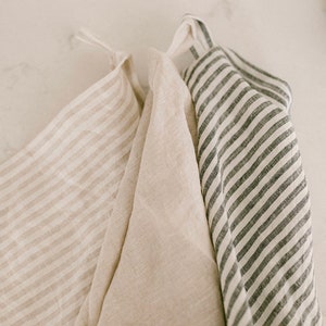 Pu&ograve; includere: Tre strofinacci da cucina in lino dai toni neutri sono appesi su uno sfondo bianco. Uno strofinaccio ha strisce beige, un altro &egrave; beige tinta unita e il terzo ha strisce grigio scuro. Gli strofinacci sono piegati e hanno anelli per appenderli.
