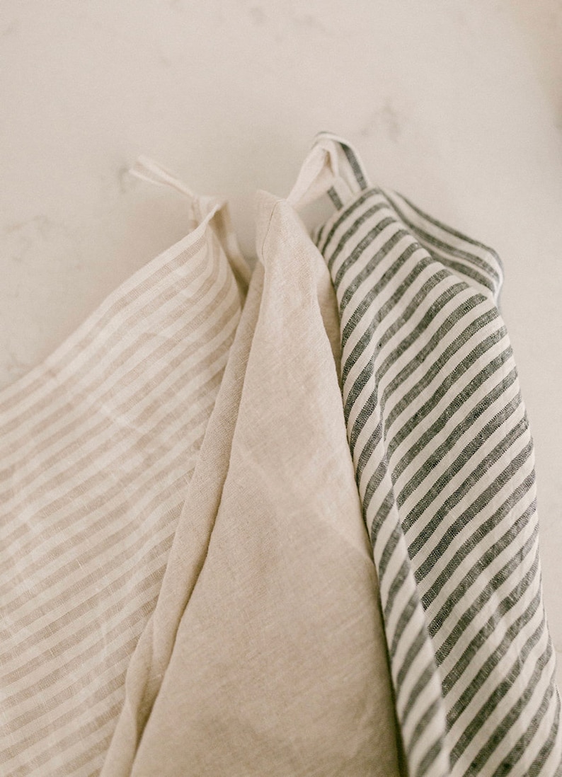 Pu&ograve; includere: Tre strofinacci da cucina in lino dai toni neutri sono appesi su uno sfondo bianco. Uno strofinaccio ha strisce beige, un altro &egrave; beige tinta unita e il terzo ha strisce grigio scuro. Gli strofinacci sono piegati e hanno anelli per appenderli.