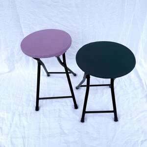May include: Two folding stools with round seats. One stool has a lavender seat, the other a dark teal seat. Both have black metal frames and legs. The stools are set against a white background.