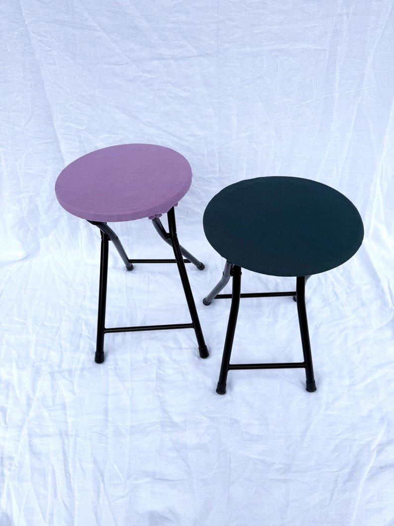 May include: Two folding stools with round seats. One stool has a lavender seat, the other a dark teal seat. Both have black metal frames and legs. The stools are set against a white background.