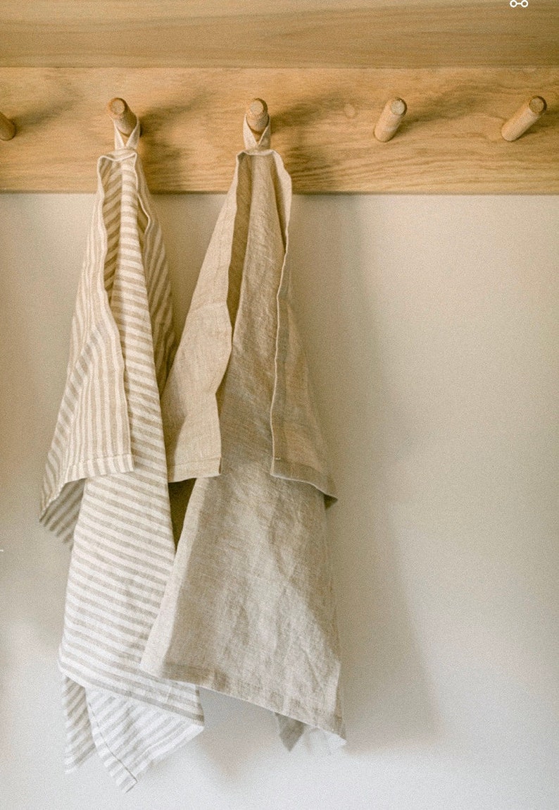 Pu&ograve; includere: Due canovacci di lino appesi a pioli di legno. Un asciugamano &egrave; beige tinta unita, l'altro &egrave; beige con strisce bianche verticali. Gli asciugamani sono piegati e drappeggiati, mostrando la loro consistenza e la tavolozza di colori neutri.