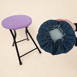 以下が含まれることがあります： 丸いラベンダー色の座面と黒い金属製の脚が付いた折りたたみスツール。明るい青色の内側が付いた濃い青色のギャザー生地のカバーが手に持たれています。カバーはスツール用であるようです。