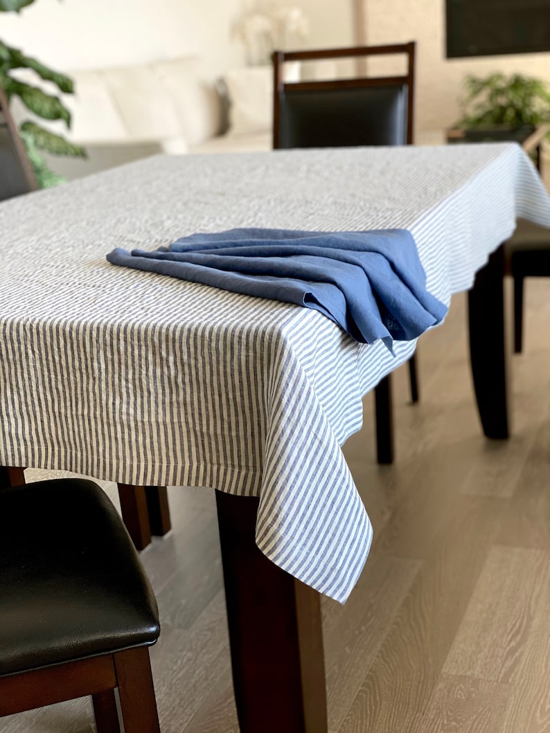 May include: A white linen tablecloth with blue stripes, folded in half and placed on a wooden table. A blue linen napkin is folded on top of the tablecloth.