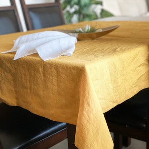 May include: A yellow linen tablecloth draped over a wooden table with two chairs. The tablecloth has a natural, slightly wrinkled texture. A white linen napkin is folded on the table.