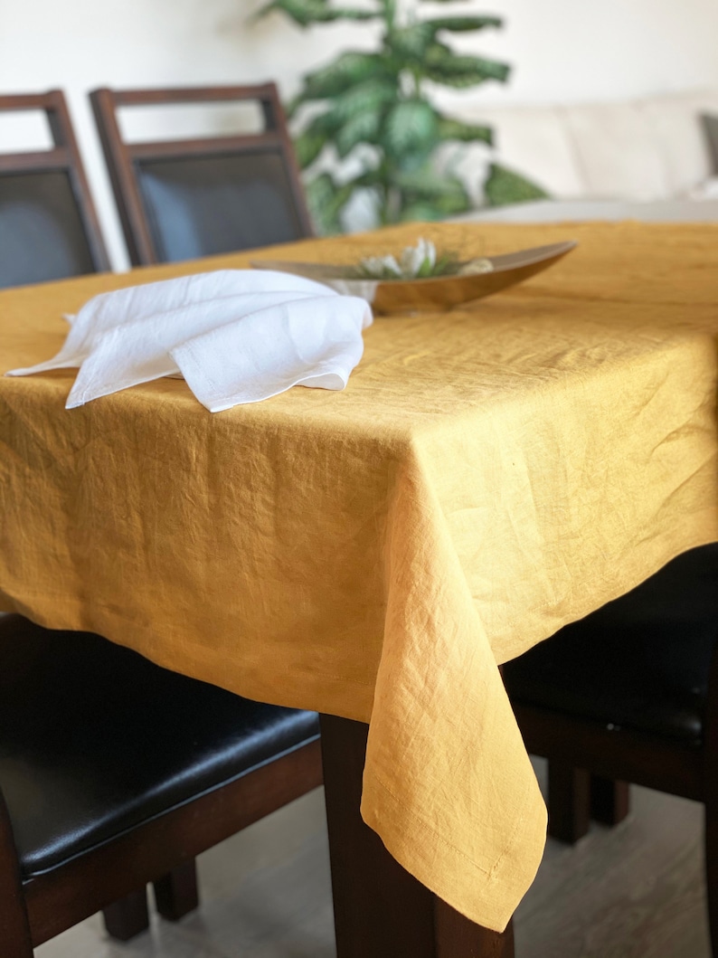 May include: A yellow linen tablecloth draped over a wooden table with two chairs. The tablecloth has a natural, slightly wrinkled texture. A white linen napkin is folded on the table.