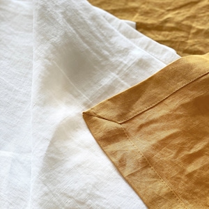 May include: Close-up of a white linen sheet folded over a mustard yellow linen sheet. The sheets are textured and have a subtle sheen.