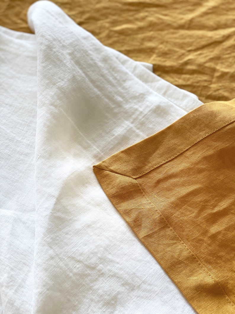 May include: Close-up of a white linen sheet folded over a mustard yellow linen sheet. The sheets are textured and have a subtle sheen.