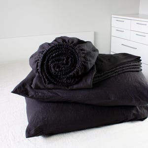 May include: A set of black linen bedding, including a fitted sheet, flat sheet, and pillowcases. The bedding is folded and arranged on a white bed.