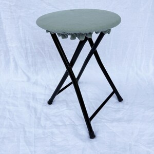 May include: A folding stool with a round, sage green seat and black metal legs. The seat has a ruffled edge. The stool is set against a white background.