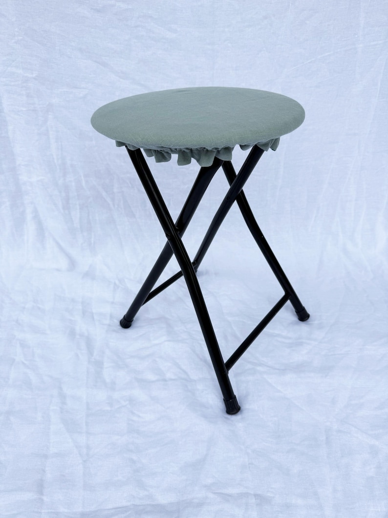 May include: A folding stool with a round, sage green seat and black metal legs. The seat has a ruffled edge. The stool is set against a white background.