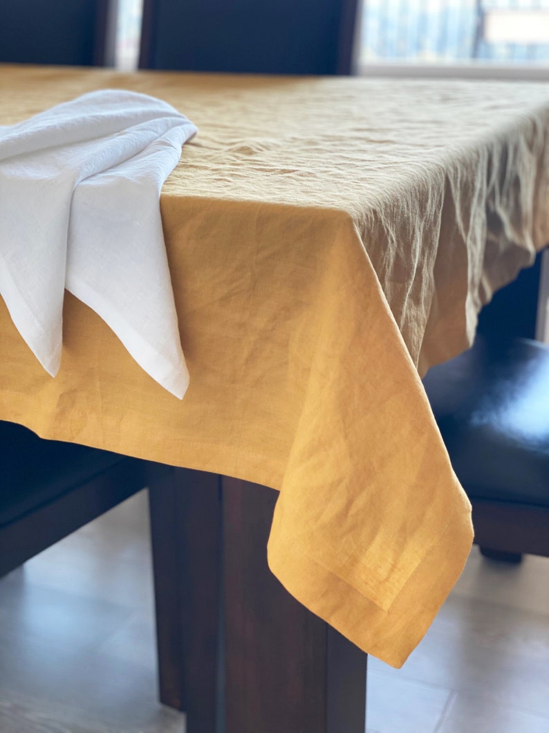 May include: A mustard yellow linen tablecloth draped over a wooden table with a white napkin folded on top.
