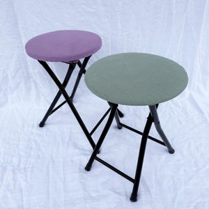 May include: Two folding stools with round seats. One stool has a purple seat, the other has a green seat. Both stools have black metal frames and legs. The stools are on a white background.