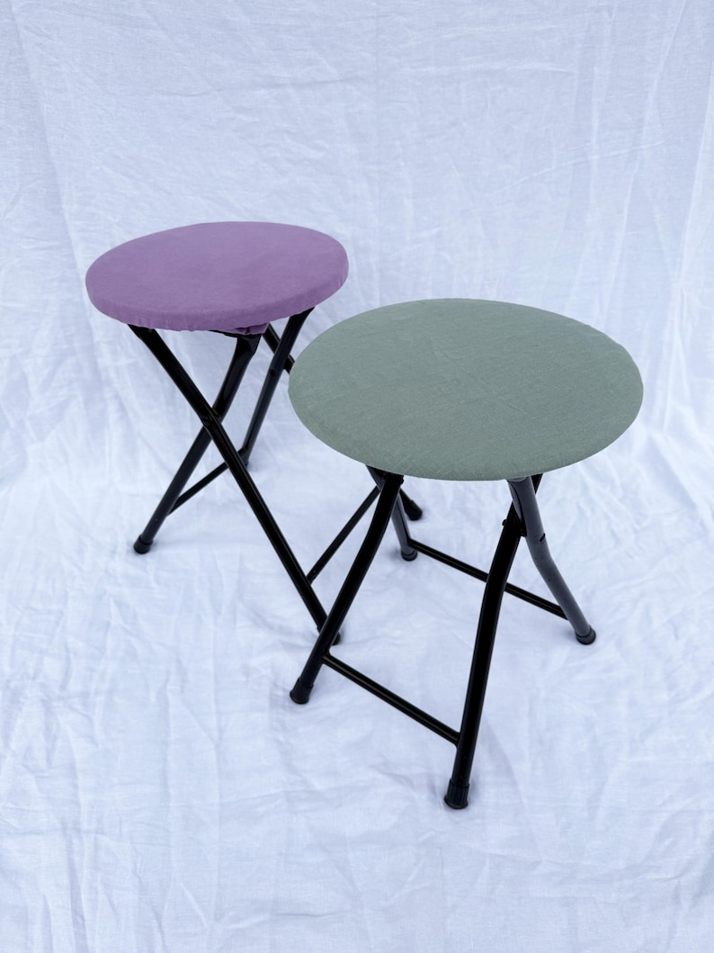 May include: Two folding stools with round seats. One stool has a purple seat, the other has a green seat. Both stools have black metal frames and legs. The stools are on a white background.