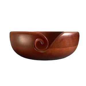 May include: A wooden yarn bowl with a rich, dark brown finish. The bowl has a curved shape with a spiral design on the side for holding yarn. The bowl is designed to keep yarn from rolling away while knitting or crocheting.