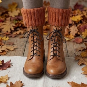 May include: A pair of tan leather lace-up boots featuring dark brown laces and thick, ribbed, burnt orange knit boot cuffs that extend above the ankle. The boots have a rounded toe, a sturdy sole, and a smooth finish, worn over grey leggings.