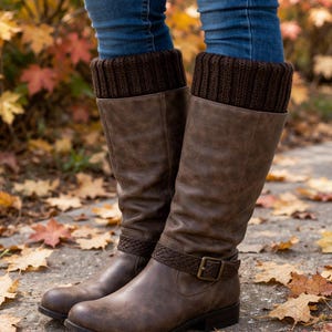 May include: Brown knee-high boots with a buckle and braided strap detail. The boots are paired with brown knit boot cuffs and blue jeans. The background features fall foliage and fallen leaves.