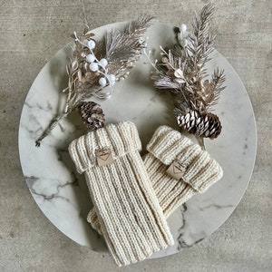 May include: A pair of cream-colored, ribbed knit leg warmers with a folded cuff, displayed on a marble-patterned plate with decorative silver and white floral accents and pinecones. Each warmer has a small tag.