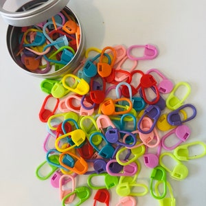 May include: A tin container filled with colorful plastic stitch markers. The markers are in various shapes and sizes, including circles, ovals, and squares. The markers are scattered around the container, creating a vibrant and playful display.