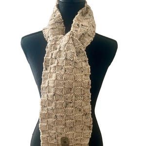 May include: A beige, hand-knitted scarf with a textured, checkered pattern. The scarf is displayed on a black mannequin and features a small, rectangular tag. The scarf is made of a soft, speckled yarn.