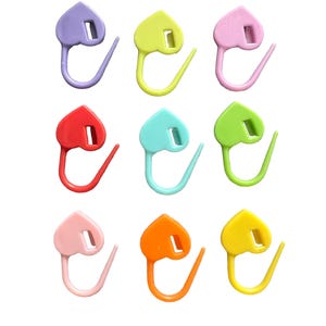 May include: A set of nine heart-shaped plastic stitch markers arranged in a three-by-three grid. The markers are colored in shades of purple, yellow, pink, red, turquoise, lime green, light pink, orange, and bright yellow, each with a small rectangular slot.