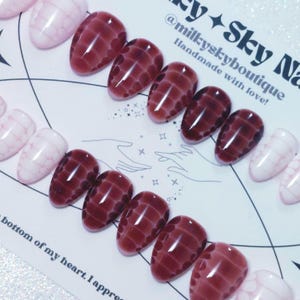 May include: Oval-shaped false nails in a burgundy and pink color scheme. The burgundy nails feature a textured pattern, while the pink nails have a subtle grid design. The image includes text that reads "@milkyskyboutique Handmade with love!"