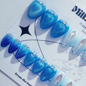 May include: A set of blue and white artificial nails with a glossy finish. Some nails feature a gradient effect, while others have sparkling embellishments. The nails are arranged on a white surface with text that reads "Milk".