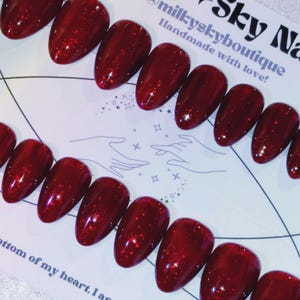 May include: A set of glossy, oval-shaped, red press-on nails with glitter accents. The nails are arranged on a white surface with the text "milkyskynails" and "Handmade with love!" printed on the top right corner.