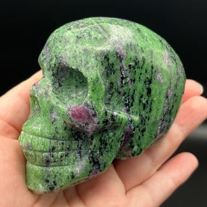 May include: A polished skull carving crafted from green zoisite with prominent inclusions of deep magenta ruby and black hornblende. The stone features a smooth, glossy surface with a mottled texture, showcasing the natural color variations.