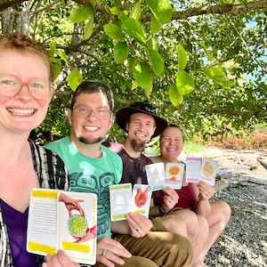 May include: Four people are sitting under a tree on a beach, holding colorful cards with illustrations and text. The cards have titles like "Garden Seeds" and "Strategies".