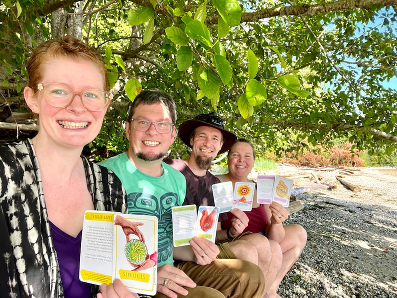 May include: Four people are sitting under a tree on a beach, holding colorful cards with illustrations and text. The cards have titles like "Garden Seeds" and "Strategies".
